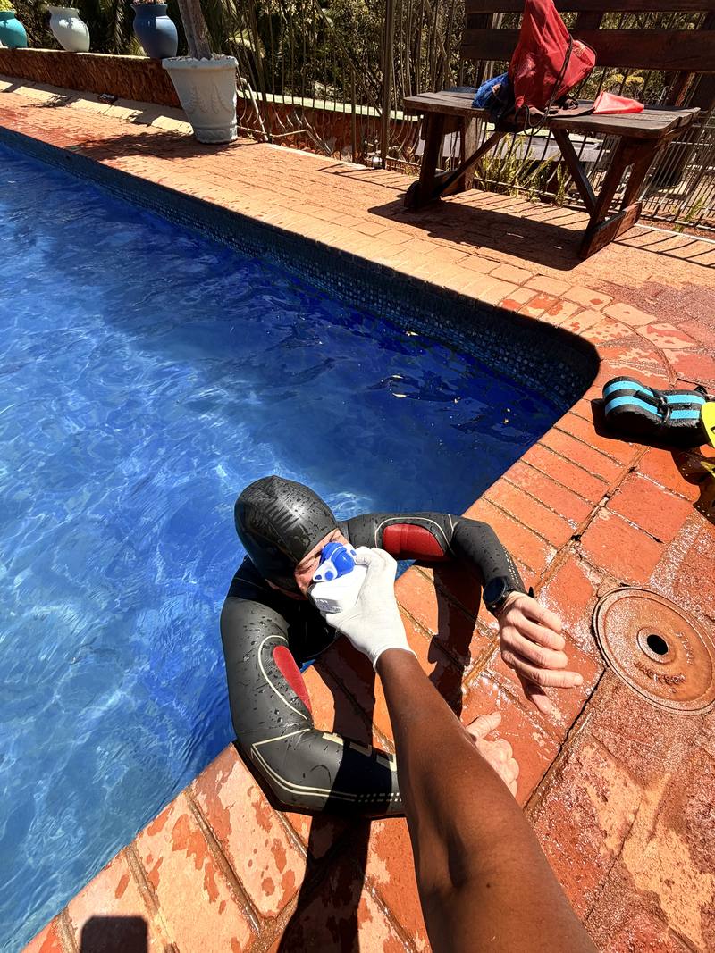 An athlete in a wetsuit at the pool edge wearing a performance testing mask while a coach assists